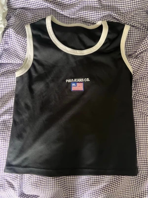 Svart linnetopp från Polo Jeans Co. - Snygg svart linnetopp från Polo Jeans Co. med vita kontrastsömmar runt hals och ärmhål. Broderad logga och amerikansk flagga på bröstet. Perfekt till jeans eller kjol för en casual look.