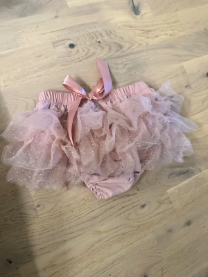 Rosa volangtrosor med glitter och rosett - Supersöta rosa trosor med flera lager volang i tyll och glittriga detaljer. Trosorna har en bred resår i midjan och en dekorativ satinrosett framtill. Perfekta för dig som gillar en lekfull och gullig stil.Rosa volangtrosor med glitter och rosett