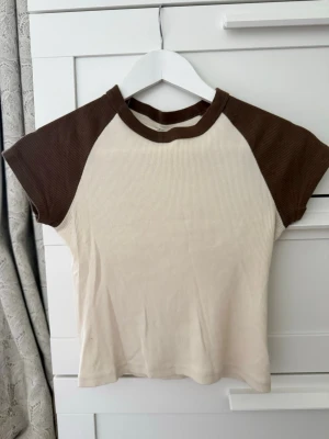 Beige och brun ribbad t-shirt - Säljer en beige t-shirt med bruna kontrastärmar och ribbad struktur. T-shirten har korta ärmar och en rund halsringning med brun kant. Perfekt för en avslappnad och trendig look.