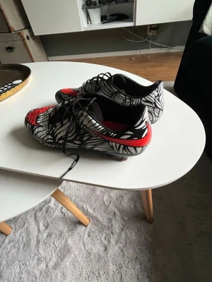 Svarta och vita hypervenoms  - Snygga hypervenoms med svartvitt grafiskt mönster och röda detaljer framtill och på hälen. Skorna har snörning och låg profil, perfekt för dig som vill sticka ut på planen. Yttermaterialet ser ut att vara syntetiskt för bra grepp och kontroll.