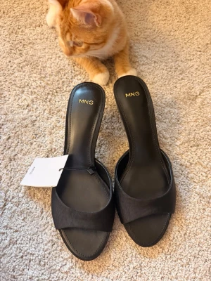 Svarta klackar från mango - Stilrena svarta slip-in pumps från MNG med öppen tå och bred rem över foten. Skorna har en elegant, smal klack och är tillverkade i ett slätt material med diskret struktur. Perfekta för att lyfta din outfit med en klassisk touch.