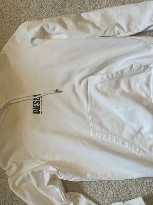 Vit hoodie från Diesel med logga - Säljer en clean vit hoodie från Diesel med svart logga på bröstet. Hoodien har en stor magficka, dragsko i huvan och är gjord i mjukt bomullsmaterial. Perfekt för dig som gillar enkel och stilren streetwear.