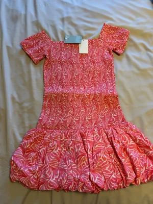  Ny trendig Rosa kortklänning med volang Stl S/M  - Supersöt kortklänning i rosa och vitt med ett livligt bladmönster. Klänningen har korta ärmar, offshoulder-skärning och en bred volang längst ner. Tillverkad i ett lätt och mjukt material, perfekt för varma dagar. Märke syns på lappen men är otydligt.