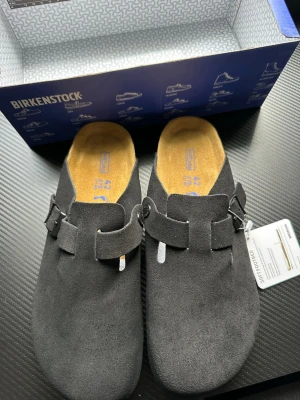 Svarta Birkenstock tofflor i mocka - Säljer ett par svarta Birkenstock tofflor i mjuk mocka med justerbart spänne och klassisk korksula. Tofflorna har öppen häl och bekväm innersula i mocka. Perfekta för chill dagar hemma eller när du vill ha det extra bekvämt.