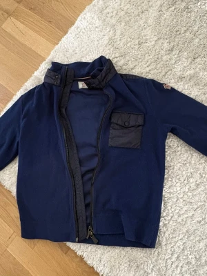 Mörkblå moncler fleece - Mörkblå jacka med hög krage och dragkedja framtill. Jackan har en bröstficka med lock och knapp samt detaljer i ett annat tyg på fickan och kragen. Perfekt för dig som gillar en stilren och sportig look.