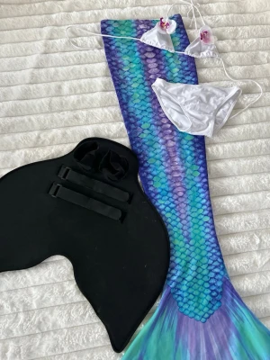 AbyssMermaid sjöjungfrudräkt set S - Säljer ett färgglatt sjöjungfrudräkt set från Abyss mermaid i storlek S men tyget är stretchigt. Bikinin är i storlek 12 år. Setet inkluderar en sjöjungfrustjärt i blå, lila och turkosa toner med fiskfjällsmönster, vit bikini med snörning och blommor samt en svart monofena. Materialet är stretchigt och snabbtorkande.