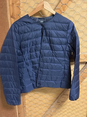 Blå dunjacka från Uniqlo - Säljer en blå, tunn och lätt dunjacka från Uniqlo i storlek M. Jackan har rund halsringning, raka ärmar och diskreta knappar framtill. Perfekt för lager-på-lager och enkel att packa ner i väskan 🌸
