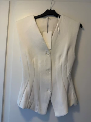 Vit väst från Zara med knappar och v-ringning - Stilren vit väst med v-ringad hals och tre knappar framtill. Västen har en figurnära passform och är tillverkad i ett mjukt tyg som ger en elegant look. Perfekt att matcha med skjorta eller t-shirt för en trendig outfit.