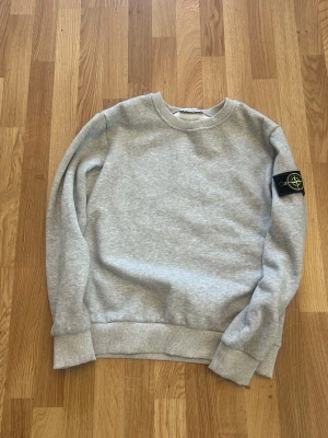 Grå sweatshirt från Stone Island - Säljer en stilren grå sweatshirt från Stone Island med klassisk rund hals och ribbade muddar. Tröjan har den ikoniska svarta patchen med logga på vänster ärm. Perfekt för en avslappnad och trendig look.
