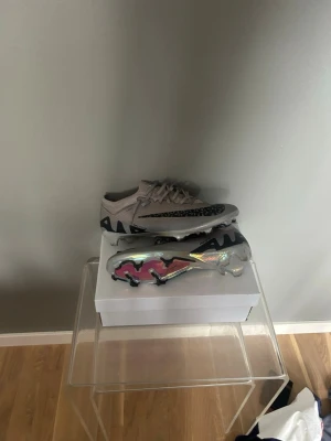 Nike Mercurial Zoom fotbollsskor - Säljer ett par Nike Mercurial Zoom fotbollsskor i ljusgrått med svarta detaljer och mönster. Skorna har en futuristisk, iriserande sula med rosa inslag och tydliga dobbar för bra grepp på planen. Snygg och modern design för dig som vill sticka ut på fotbollsplanen.  Skriv om ni har några tankar 