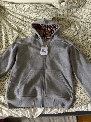 Grå hoodie från Burberry med dragkedja - Säljer en grå hoodie från Burberry med dragkedja och klassiskt rutigt foder i huvan. Hoodien har två stora fickor framtill och ribbade muddar vid ärmslut och nederkant. Perfekt för dig som gillar stilrena och exklusiva plagg.