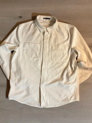 Vit overshirt från ARKET - Stilren vit overshirt från Zara i bomull med två bröstfickor och klassisk krage. Skjortan har knappar framtill och långa ärmar, perfekt för lager på lager och enkel att matcha med olika outfits.