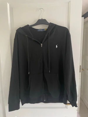 Polo Ralph Lauren - Svart hoodie med dragkedja från Polo Ralph Lauren. Klassisk design med huva, dragsnören och fickor framtill. Liten vit logga broderad på bröstet. Knappt använd då den var lite för liten. Det är storlek XL men skulle säga att den passar som L.