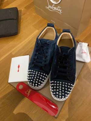 Mörkblå Louboutin sneakers med nitar - Snygga mörkblå sneakers från Christian Louboutin i mocka med silverfärgade nitar på tån. Skorna har klassisk låg modell, svarta skosnören och beige innerfoder. Perfekta för dig som vill sticka ut med exklusiv design och edgy detaljer.