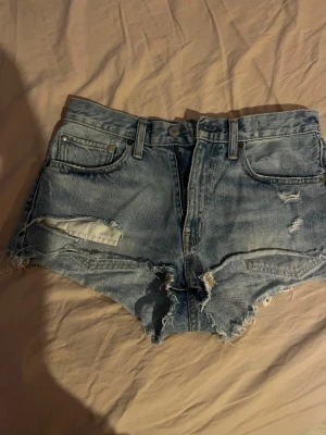 Blåa högmidjade jeansshorts - Säljer ett par blåa högmidjade jeansshorts från Ralph Lauren med slitna detaljer och råa kanter. Klassisk femficksmodell med bälteshällor och knappgylf. Perfekta för sommaren och ger en avslappnad vibe. W:28