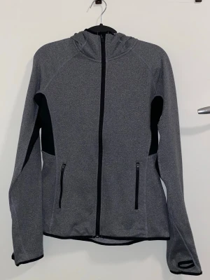 Grå sporthoodie med svarta detaljer - Snygg grå sporthoodie med svarta detaljer och dragkedja framtill. Tröjan har två fickor med dragkedja, huva och tumhål vid ärmsluten. Perfekt för träning eller löpning. Tillverkad i ett stretchigt funktionsmaterial som andas.