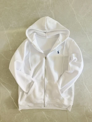 Vit hoodie med dragkedja från Polo Ralph Lauren - Snygg vit hoodie från Polo Ralph Lauren med dragkedja framtill och klassisk blå logga broderad på bröstet. Hoodien har huva med dragsko och två fickor framtill. Tillverkad i mjukt och bekvämt material, perfekt för en chill och stilren look.
