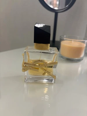 Yves Saint Laurent Libre parfym - Säljer en elegant parfym från Yves Saint Laurent, Libre. Flaskan är fyrkantig i genomskinligt glas med guldfärgad YSL-logga och detaljer. Svart asymmetrisk kork och guldmönstrad hals ger en lyxig känsla. Innehållet är ljust bärnstensfärgat.