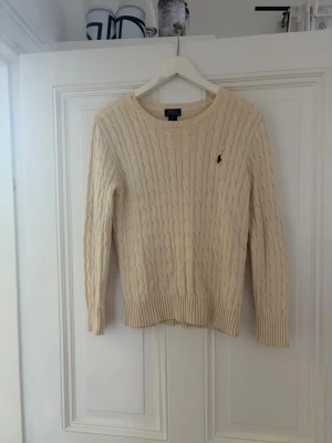 Kabelstickad beige tröja från Ralph Lauren - Kabelstickad beige tröja från Ralph Lauren med rund halsringning och diskret broderad logga på bröstet. Tröjan har långa ärmar och ribbade muddar vid ärmslut och nederkant. Perfekt för en stilren och klassisk look.