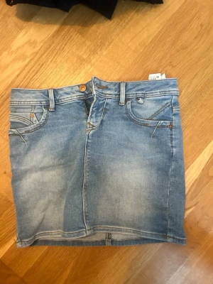 Blå jeanskjol från LTB - Säljer en klassisk blå jeanskjol från LTB med fem fickor, bälteshällor och knappstängning fram. Kjolen har en kort passform och är gjord i mjukt denimtyg. Perfekt till sneakers eller boots för en avslappnad look.