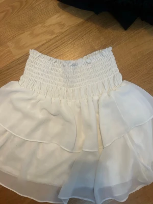 Vita skorts med volanger - Säljer ett par vita skorts med bred smockresår i midjan och dubbla lager volanger i ett tunt, flowy tyg. Perfekta för en trendig och luftig look. Passar till crop tops och sneakers för en avslappnad stil.