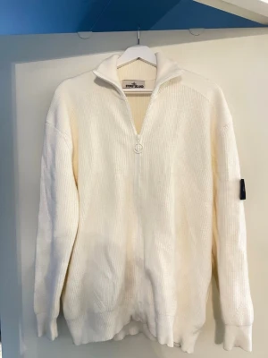 Vit stickad ziptröja från Stone Island - Vit stickad tröja från Stone Island med dragkedja framtill och hög krage. Tröjan har ribbstickad struktur och den klassiska Stone Island-loggan på vänster ärm. Perfekt för lager-på-lager och snygg till jeans.