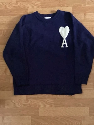 Mörkblå stickad tröja - Mörkblå stickad tröja från  med ett vitt hjärta och bokstaven 'A' broderat på bröstet. Tröjan har rund halsringning, långa ärmar och ribbade muddar vid ärmslut och nederkant. Perfekt för en trendig och avslappnad look.