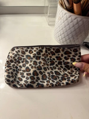 Leopardmönstrad sminkväska - Snygg liten sminkväska med leopardmönster i beige, brunt och svart. Väskan har dragkedja upptill och är fodrad med matchande leopardmönster även på insidan. Perfekt för att förvara smink eller småprylar i väskan.