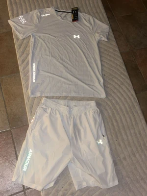 Grått träningsset från Under Armour - Säljer ett grått träningsset från Under Armour med t-shirt och shorts. Setet har tryckta loggor och textdetaljer i vitt och svart. Tillverkat i ett lätt och snabbtorkande material som passar perfekt för träning och sportaktiviteter.