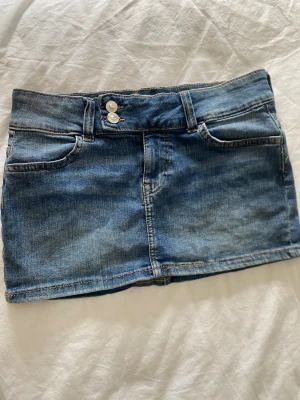  jeanskjol  - Snygg blå jeanskjol i kort modell med klassiska fickor fram och bak samt dubbla silverknappar i midjan. Kjolen har en rak passform och slitna detaljer för en avslappnad look. Perfekt till vardags med en t-shirt eller hoodie.