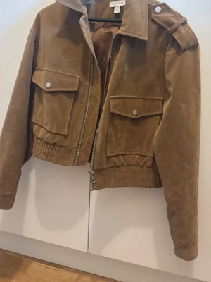 Brun skinnjacka med fickor - Snygg brun skinnjacka från H&M med två stora fickor framtill, silverfärgade knappar och dragkedja. Jackan har resår i midjan och axelklaffar för en cool look. Perfekt för dig som vill ha en trendig och stilren jacka i garderoben.