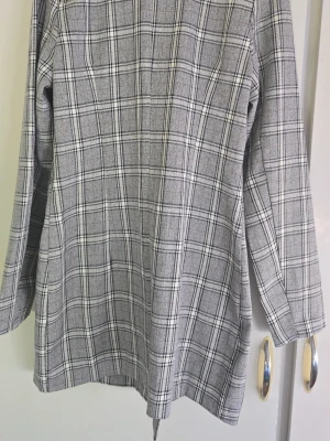 Grå rutig dubbelknäppt rock - Snygg grå rock med svartvitt rutmönster och dubbelknäppning framtill. Klassisk krage och raka ärmar ger en clean look. Perfekt för dig som gillar stilrena och tidlösa plagg. Materialet känns mjukt och har en lätt struktur.