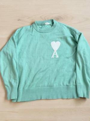 AMI Paris Ami de Coeur Sweater Mint Green Heart Logo Knit Crewneck Size M - Mintgrön stickad tröja från Ami Paris med vitt hjärta och bokstaven A på bröstet. Tröjan har ribbade muddar vid ärmslut och nederkant samt en klassisk rund halsringning. Perfekt för dig som gillar stilrena detaljer och trendiga färger.