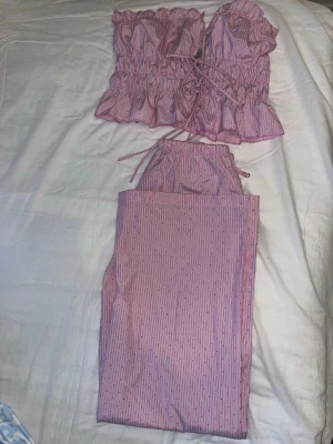 Rosa randigt set med volanger - Säljer ett tvådelat set i rosa med tunna vita diamanter. Toppen har volangkant, smock och knytning framtill. Tillhörande byxa med samma mönster och resår i midjan. Perfekt för dig som gillar söta detaljer och bekväma material. Överdelen är S o underdelen är XXS