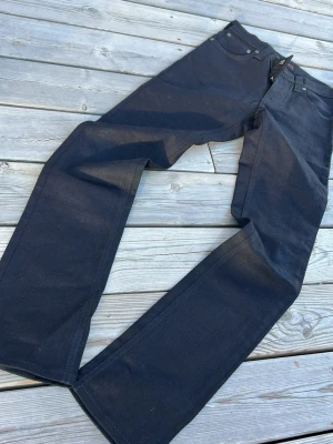 Svarta jeans från Nudie Jeans - Snygga svarta jeans från Nudie Jeans med raka ben och klassisk femficksmodell. Jeansen har knappgylf och är gjorda i ett kraftigt denimtyg som ger en clean och stilren look. Perfekta för dig som gillar tidlös och enkel stil.