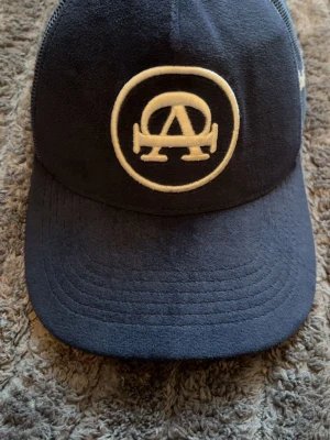 Mörkblå meshkeps med vit logga - Snygg mörkblå keps med vit broderad logga framtill. Kepsen har böjd skärm och mesh på sidorna och bak för extra ventilation. Perfekt för en avslappnad streetwear-look. Justerbar baktill för bästa passform.