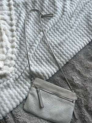 Ljusgrå axelväska med mönster - Snygg ljusgrå axelväska i konstläder med justerbar rem. Väskan har ett dekorativt blommigt mönster på framsidan och två dragkedjefack, perfekt för mobilen och småsaker. Enkel och stilren design som passar till det mesta.