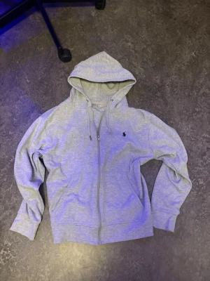Grå zip hoodie från Polo Ralph Lauren - Snygg gråmelerad hoodie med dragkedja från Polo Ralph Lauren. Klassisk huva med dragsko och den ikoniska lilla loggan broderad på bröstet. Mjuk bomullsblandning och två fickor framtill. Perfekt för en chill och avslappnad stil.