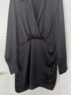 Svart omlottklänning från Zara storlek M - Stilren svart kortklänning med omlott-design och långa ärmar. Klänningen har en lätt glansig yta och breda manschetter vid ärmsluten. Perfekt för dig som gillar en enkel men elegant look.