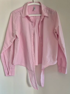 Ljusrosa skjorta/knytblus - Snygg ljusrosa skjorta med långa ärmar och knyt framtill. Skjortan har klassisk krage och knappar hela vägen. Perfekt för en trendig och avslappnad look. Tillverkad i mjuk bomull som känns skön mot huden.