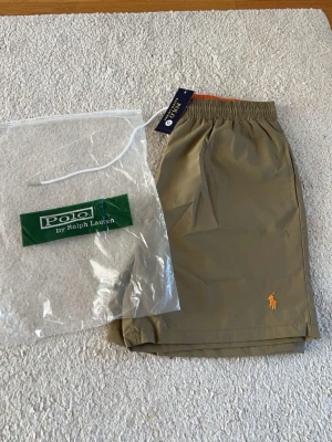 bad byxor från Polo Ralph Lauren XL passar L - Snygga beige shorts från Polo Ralph Lauren med klassisk orange broderad logga framtill. Shortsen har elastisk midja och är tillverkade i ett lätt och bekvämt material, perfekta för varma dagar.