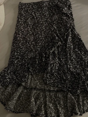 Svart blommig långkjol med volang - (Känns som smal) Svart långkjol med vita små blommor och volangkant nedtill. Kjolen har en flowy passform och är gjord i ett lätt material som känns skönt mot huden. Perfekt för dig som gillar en romantisk och trendig stil.