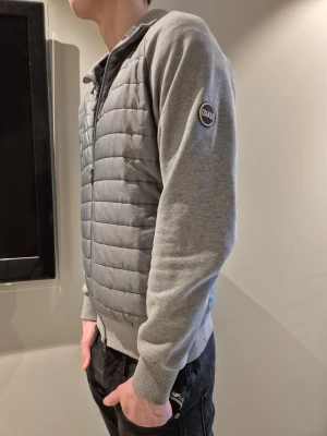 Grå ziptröja från Colmar - Snygg grå ziptröja från Colmar med quiltad front och mjuka ärmar. Tröjan har ribbade muddar, hög krage och en diskret logga på ärmen. Tveka inte att höra av er vid frågor.