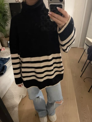 Randig stickad polotröja från COS - Säljer en svart och beige stickad polotröja från COS med breda ränder nertill och på ärmarna. Tröjan har oversized passform, lång ärm och hög krage. Perfekt för en chill och stilren look.