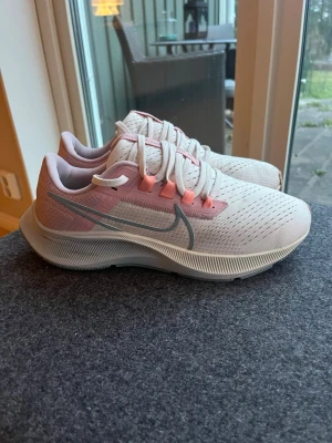 Nike Air Zoom Pegasus - Nike Air Zoom Pegasus sneakers i ljusrosa och vitt med snygga detaljer i rosa och silver. Skorna har snörning, ventilerande meshmaterial och en platt sula. Perfekta för dig som gillar sportig och fräsch stil.