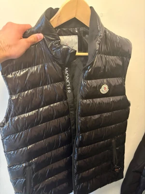 Svart dunväst från Moncler - Snygg svart dunväst från Moncler med glansig finish och klassisk logga på bröstet. Västen har ståkrage, dragkedja framtill och två fickor med dragkedja. Perfekt för lager-på-lager och streetstyle.
