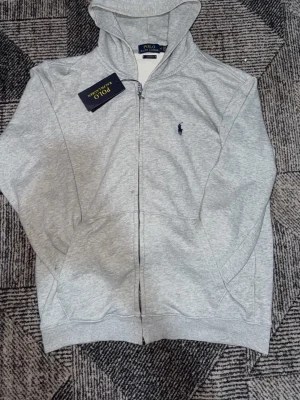 Grå hoodie från Polo Ralph Lauren - Snygg grå hoodie från Polo Ralph Lauren med dragkedja framtill och klassisk blå logga broderad på bröstet. Hoodien har huva, ribbade muddar och fickor på sidorna. Tillverkad i mjukt bomullsmaterial som är skönt att bära. Tag medföljer EJ lite skador 