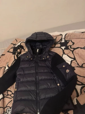 Svart pufferjacka med huva från Moncler - Snygg svart pufferjacka från Moncler med stickade ärmar och huva. Jackan har dragkedja framtill, två fickor med tryckknappar och en Moncler-logga på ärmen. Perfekt för kalla vinterdagar och riktigt trendig look.