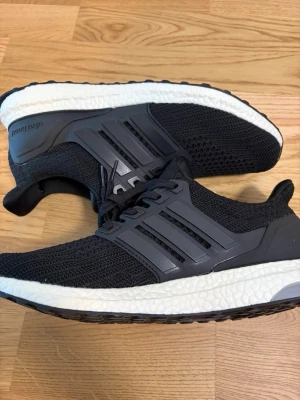 Svarta Adidas Ultraboost sneakers - Svarta Adidas Ultraboost sneakers med stickad ovandel i mesh och klassiska tre ränder på sidan. Skorna har en vit, responsiv Boost-sula och svarta detaljer. Perfekt för dig som gillar sportig och clean stil. Lågt skaft och snörning för skön passform.