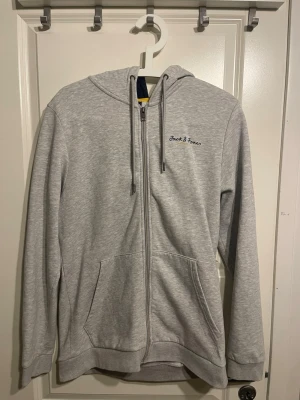 Ljusgrå hoodie från Jack & Jones - Ljusgrå zip-hoodie från Jack & Jones med broderad logga på bröstet. Hoodien har dragsko i huvan, kängurufickor och ribbade muddar. Insidan av huvan är mörkblå med en gul detalj. Perfekt för en avslappnad stil.